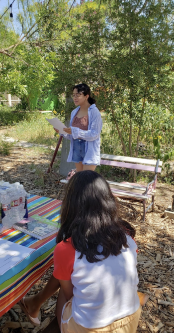 An ABC student in a floral blouse and jean shorts reads her poetry outside in front of a wooden bench while other students sit at a picnic table with their own notebooks and snacks.